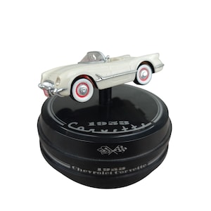 May include: A white 1953 Chevrolet Corvette model car with red-trimmed wheels and a silver trim. The car sits on a black base with the words "1953 Corvette" and a checkered flag emblem.