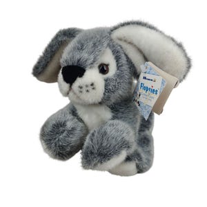 May include: A plush gray and white bunny rabbit with long floppy ears. The stuffed animal has a black nose and brown eyes. A tag attached to the ear reads "Flopsies Collection".