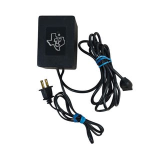 May include: A black rectangular power adapter featuring a silver Texas Instruments logo on the front. The device includes a two-prong electrical plug and a long black cord secured with blue rubber bands, ending in a small rectangular connector.