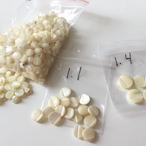 Peut inclure: Plusieurs petits boutons ronds blanc cassé dans des sacs en plastique transparent. Certains sacs sont numérotés. Les boutons ont un éclat subtil et un design plat, légèrement concave, avec une seule rainure au centre. Idéal pour la couture et les projets d'artisanat.