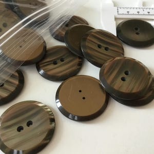 Może przedstawiać: Kolekcja dużych, okrągłych guzików w odcieniach brązu i beżu, ze wzorem w wiry. Każdy guzik ma dwa otwory do przyszywania. Niektóre guziki są w przezroczystym plastikowym woreczku, a w tle znajduje się narzędzie pomiarowe.