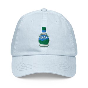 May include: Light blue baseball cap with an embroidered image of a bottle of ranch dressing. The bottle is blue and green with the word "RANCH" printed on it.
