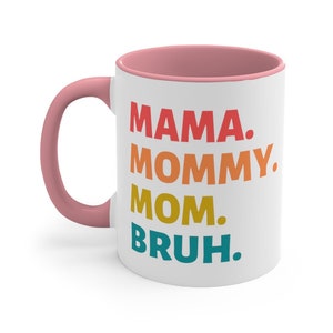 May include: White ceramic coffee mug with a pink rim and handle. The mug features a colourful graphic that reads "MAMA. MOMMY. MOM. BRUH."