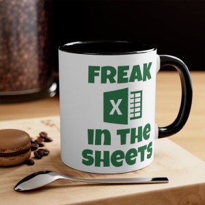 May include: White coffee mug with a black rim and handle. The mug has a green graphic of the Microsoft Excel logo and the text "FREAK in the sheets".