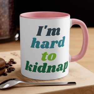 May include: White ceramic mug with a pink rim and handle. The mug features a green, blue, and black graphic that says "I'm hard to kidnap".