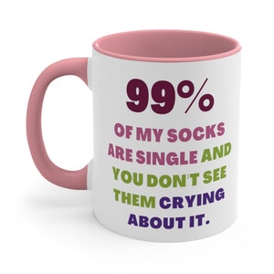 May include: White ceramic coffee mug with a pink rim. The mug has a humorous saying printed on it in colorful text: "99% of my socks are single and you don't see them crying about it."