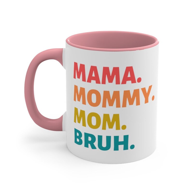 May include: White ceramic coffee mug with a pink rim and handle. The mug features a colourful graphic that reads "MAMA. MOMMY. MOM. BRUH."