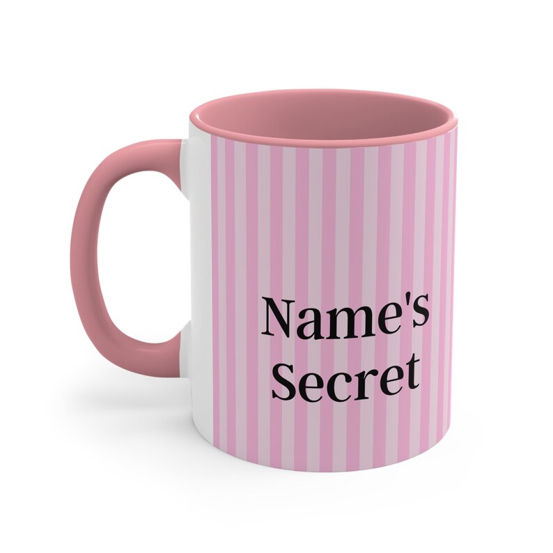 May include: A pink and white striped ceramic mug with a pink handle. The mug has the text "Name's Secret" printed on the front.