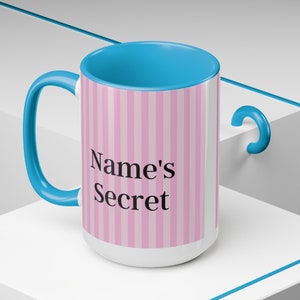 May include: A white ceramic mug with a pink and white striped design. The mug has a blue handle and a blue rim. The text "Name's Secret" is printed on the mug.
