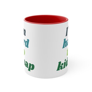 May include: White ceramic mug with a red rim. The mug has a green, blue, and black design with the text "I'd rather be napping" on the left side and "I have to kid you" on the right side.