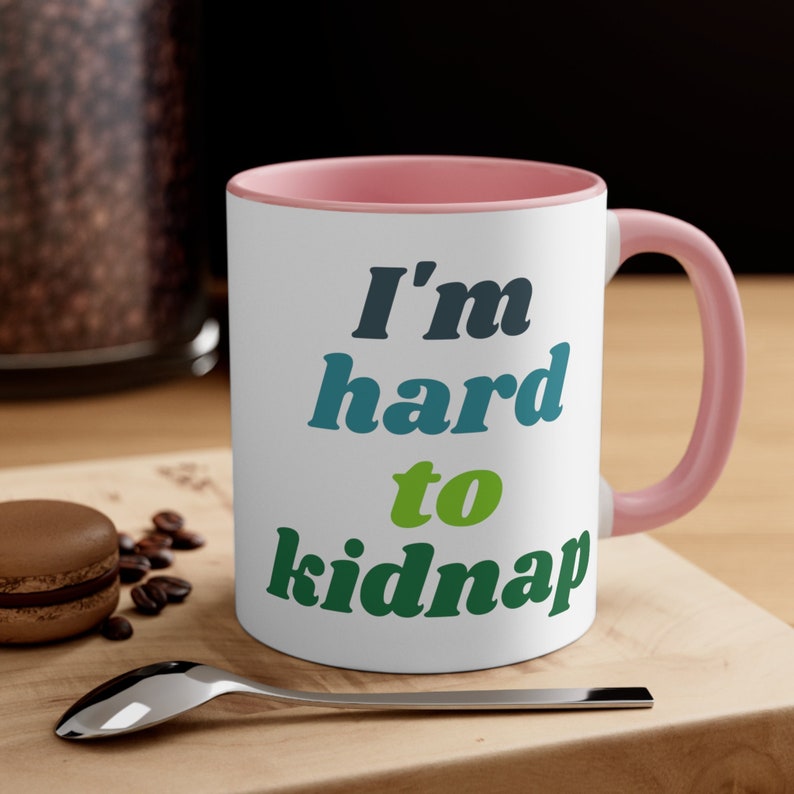 May include: White ceramic mug with a pink rim and handle. The mug features a green, blue, and black graphic that says "I'm hard to kidnap".