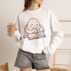 Puede incluir: Una sudadera blanca de manga larga con un gráfico de un pequeño fantasma beige, redondo y envuelto en una manta, leyendo un libro abierto con una vela pequeña y dos bloques apilados cerca, rodeado de pequeñas estrellas coloridas.