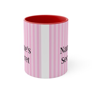 May include: A pink and white striped ceramic mug with a red rim. The mug has the text "Name's Secret" printed on it.