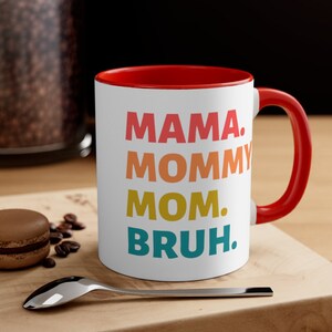 May include: White ceramic coffee mug with a red rim and handle. The mug features a colourful graphic that reads "MAMA. MOMMY MOM. BRUH."