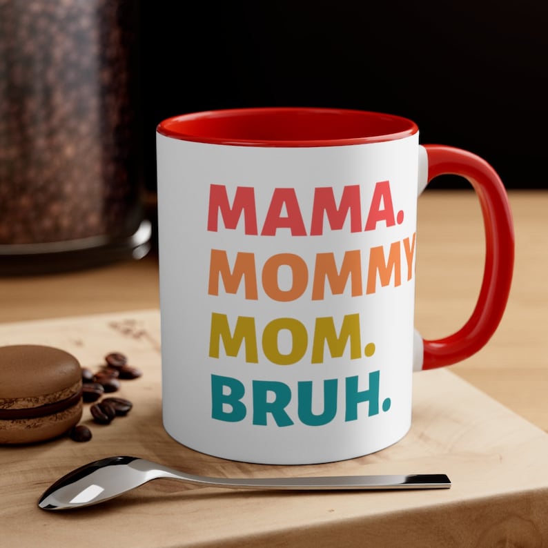 May include: White ceramic coffee mug with a red rim and handle. The mug features a colourful graphic that reads "MAMA. MOMMY MOM. BRUH."