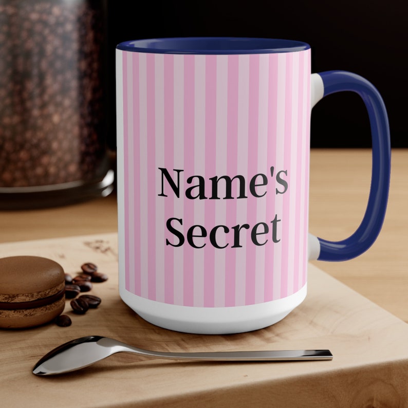 May include: White ceramic mug with a pink and white striped design and a navy blue rim. The mug has the text "Name's Secret" in black.