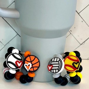 May include: Three bracelets with black beads and sports ball charms. The charms are a baseball, basketball, and volleyball. Each charm has a red heart on it.