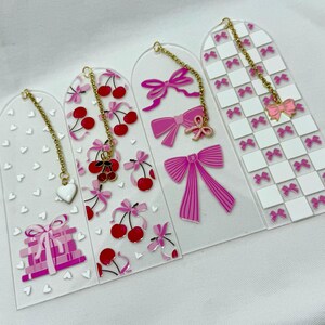 Op de afbeelding: Vier roze en witte acryl bladwijzers met gouden kettingen en bedels. De bladwijzers hebben verschillende ontwerpen, waaronder kersen, strikken, harten en een geruit patroon.