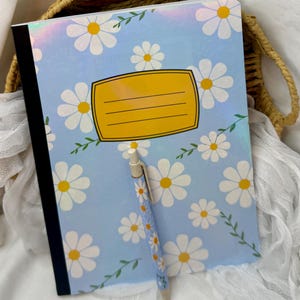 May include: A composition notebook with a light blue cover featuring white daisies with yellow centers and green leaves. A matching pen rests on the notebook. The notebook has a yellow label with lines for writing.