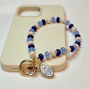 May include: A phone charm featuring a circular arrangement of alternating navy blue, light blue, and cream-colored beads. A gold-toned metal lobster clasp is attached to an oval pendant displaying a blue and white illustration of a seashell.