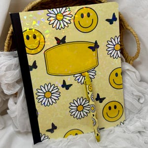 May include: A yellow notebook and pen set with a daisy, butterfly, and smiley face pattern. The notebook has a black spine and a blank yellow label. The pen has a matching design. The background is a woven basket.