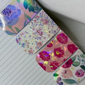 Könnte beinhalten: Vier dekorative magnetische Lesezeichen mit Blumen- und Lippendesigns. Die Lesezeichen weisen eine Vielzahl von Mustern auf, darunter rosa und lila Blumen und rosa Lippen. Die Lesezeichen haben ein holografisches, irisierendes Finish.