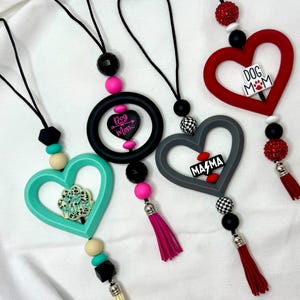May include: A collection of heart-shaped car charms in various colors, including teal, red, and gray. Each charm features a different design, such as a paw print, the words "Dog Mom," or "Mama." They are adorned with beads, tassels, and black cords.