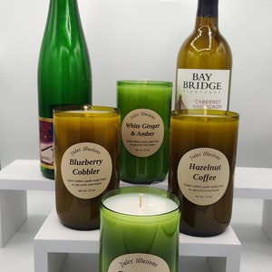 May include: Four green glass candle jars with white wax and brown labels. The labels have the text "Jules' Illusions" and the scent names: "Blueberry Cobbler", "White Ginger & Amber", "Hazelnut Coffee", and "Christmas Tree". Each candle is 12 oz. The candles are on a white shelf with two wine bottles behind them.