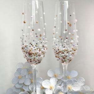 May include: Two champagne flutes adorned with pearls, iridescent beads, and gold-toned details. The decorations are densely packed at the base, thinning out towards the top. The glasses are placed on a bed of light blue artificial flowers.