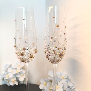 May include: Two clear champagne flutes adorned with gold, brown, and white beads and pearls. The decorative elements are clustered at the base of the glasses, creating a shimmering effect. The stems are slender and elegant, perfect for a celebration.