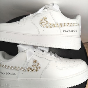May include: White sneakers with pearl and gold embellishments on the Nike swoosh. The text "08.09.2026" is on one shoe, and "Mrs. Whited" is on the other. Each shoe has a white ribbon tied at the laces.