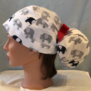 May include: A white surgical scrub hat with a grey and blue elephant print. The hat has a ponytail hole and a red bow.