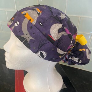 May include: A purple scrub cap with a Halloween design, featuring raccoons, bats, spiders, and mummies. It has a yellow ribbon and is designed for comfort. The fabric is patterned with various spooky elements.