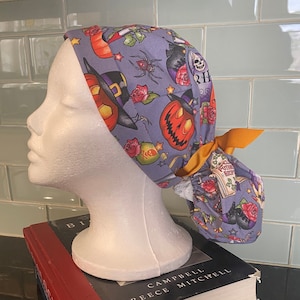 Könnte beinhalten: Eine lavendelfarbene OP-Haube mit einem Halloween-Muster mit Kürbissen, Spinnen, Hexenhüten und Rosen. Die Haube hat eine orangefarbene Schleife und wird auf einem weißen Schaufensterkopf präsentiert.