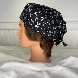 May include: A black surgical scrub cap featuring a repeating white geometric pattern. The cap is tied at the back and is displayed on a mannequin head. The background is a white wall and white fabric.