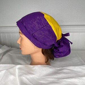 Pode incluir: Uma touca cirúrgica roxa e dourada. A touca tem as costas franzidas e é feita de tecido roxo com listras metálicas douradas e um painel amarelo na parte superior. A touca é amarrada nas costas.