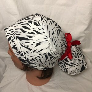 Pode incluir: Um gorro cirúrgico preto e branco estampado com um laço vermelho. O gorro tem uma abertura para rabo de cavalo na parte de trás.