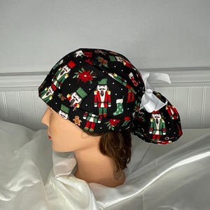 Puede incluir: Un gorro quirúrgico negro con un estampado navideño con cascanueces, flores de pascua y hombres de jengibre. El gorro está atado con un lazo blanco. El diseño incluye detalles en rojo, verde y blanco, perfecto para la temporada navideña.