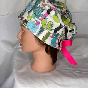 May include: A white scrub cap featuring a colorful cactus print in shades of green, blue, and pink. The cap has a bright pink ribbon and is designed for medical professionals or those in the healthcare field.