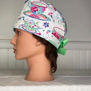 May include: White surgical scrub cap with a repeating pattern of teacups, saucers, and floral designs in pink, blue, and green. The cap has a green bow on the back. The cap is designed to be worn in a medical setting.