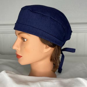 May include: A navy blue surgical scrub cap with a tie closure. The cap is made of a solid, dark blue fabric and is designed to cover the head. The tie closure allows for an adjustable fit. The cap is shown on a mannequin head.