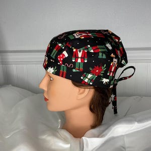 May include: A black surgical scrub cap with a festive Christmas pattern. The fabric features red, green, and white nutcrackers, stockings, and poinsettias. The cap has a tie closure at the back for an adjustable fit.