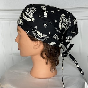 May include: Black scrub cap featuring a repeating pattern of Harry Potter-themed imagery, including the Hogwarts crest, stars, and text. The cap is tied at the back with black fabric ties. The cap is displayed on a mannequin head.
