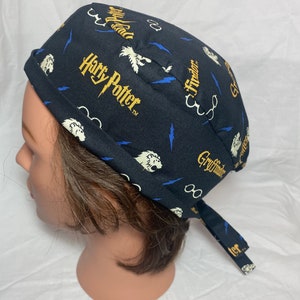 May include: Black scrub cap featuring the words "Harry Potter" in gold, along with white lion graphics, blue lightning bolts, and glasses. The cap is tied at the back and is displayed on a mannequin head.