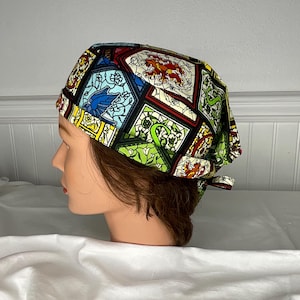 May include: A colorful surgical scrub cap featuring a stained-glass window design with birds, lions, and floral patterns. The cap is made of fabric and has a tie in the back for an adjustable fit. The color palette includes blue, green, red, and yellow.