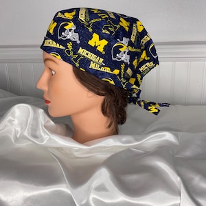 Puede incluir: Un gorro de exfoliación con estampado azul marino y amarillo con el texto "Michigan" y la letra "M", junto con cascos de fútbol. El gorro está atado en la parte posterior.