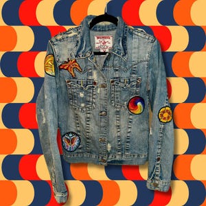 Può includere: Una giacca di jeans blu chiaro con diverse toppe ricamate colorate, tra cui una farfalla, un sole e un arcobaleno. La giacca ha un aspetto consumato ed è realizzata da True Religion.