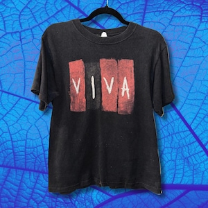 May include: Black t-shirt with the word "VIVA" in white letters, set against a backdrop of three vertical red rectangles. The shirt is hanging on a black hanger against a blue background.