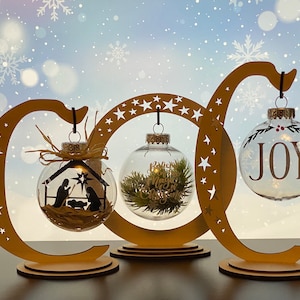 May include: Three glass ornaments on wooden stands. The ornaments are clear glass with different designs. One ornament has a nativity scene, one has a green plant, and one has the word "JOY" written on it.