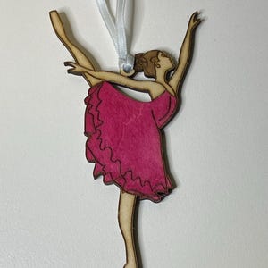 May include: A wooden ballerina ornament with a pink tutu and a white ribbon for hanging.